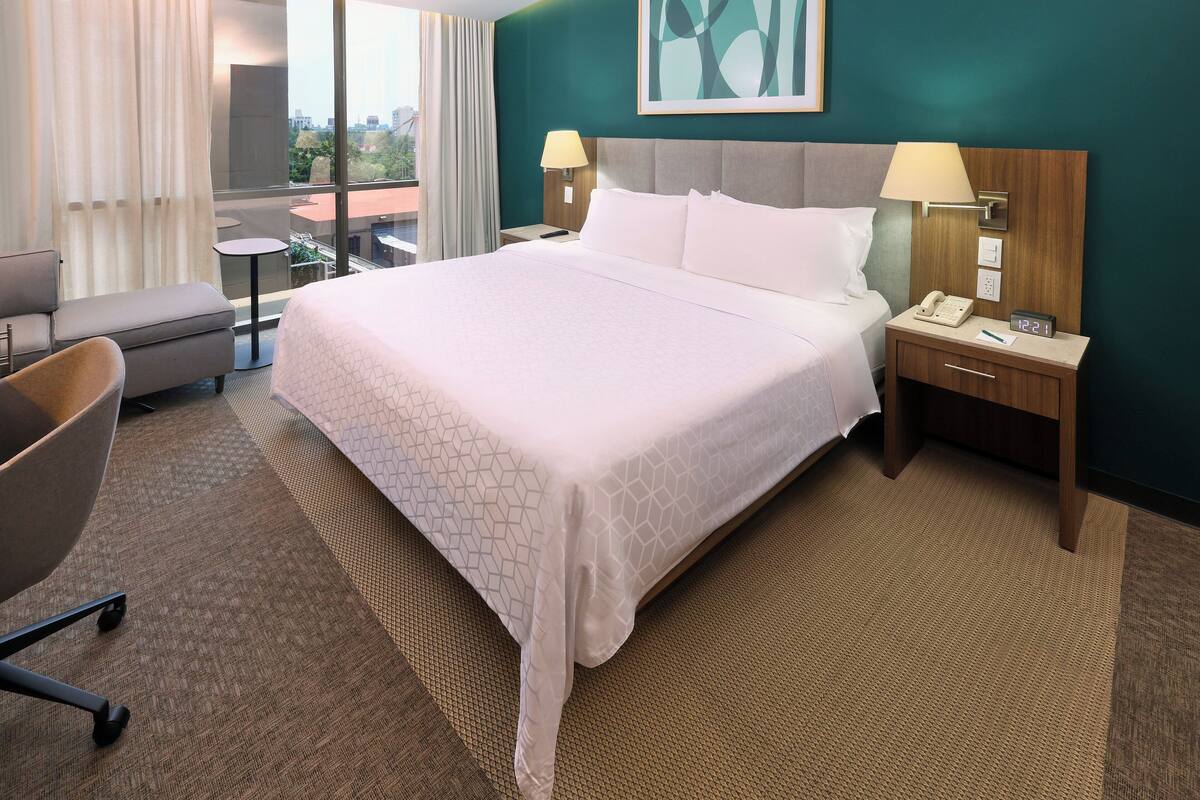 Une chambre d’hôtel avec un grand lit, une table de chevet avec une lampe, une chaise et une vue sur la ville par la fenêtre.