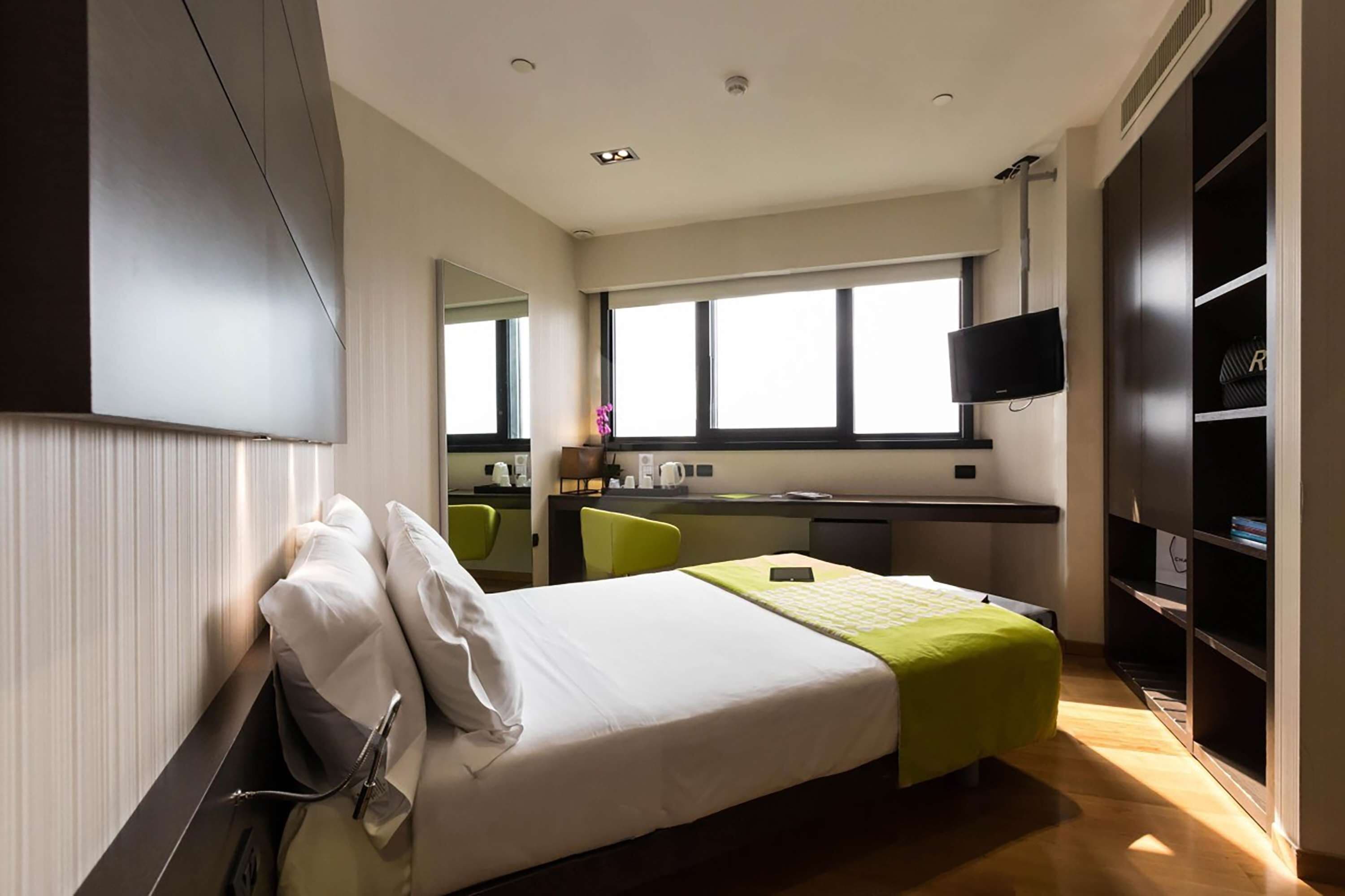 A modern hotel room with a bed, a desk, a chair, a TV, and a wardrobe.