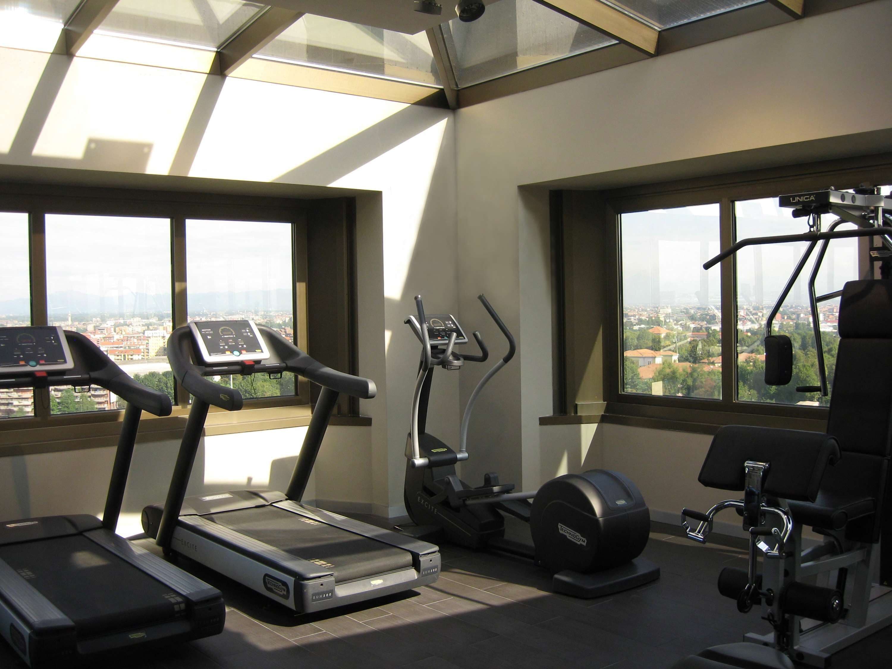 Un gimnasio con cintas de correr, elíptica y máquina de pesas, con amplias ventanas y vistas a la ciudad.