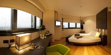 Una moderna camera d'albergo con un letto curvo, una scrivania con lampada, una sedia verde e un'ampia finestra con vista sulla città.