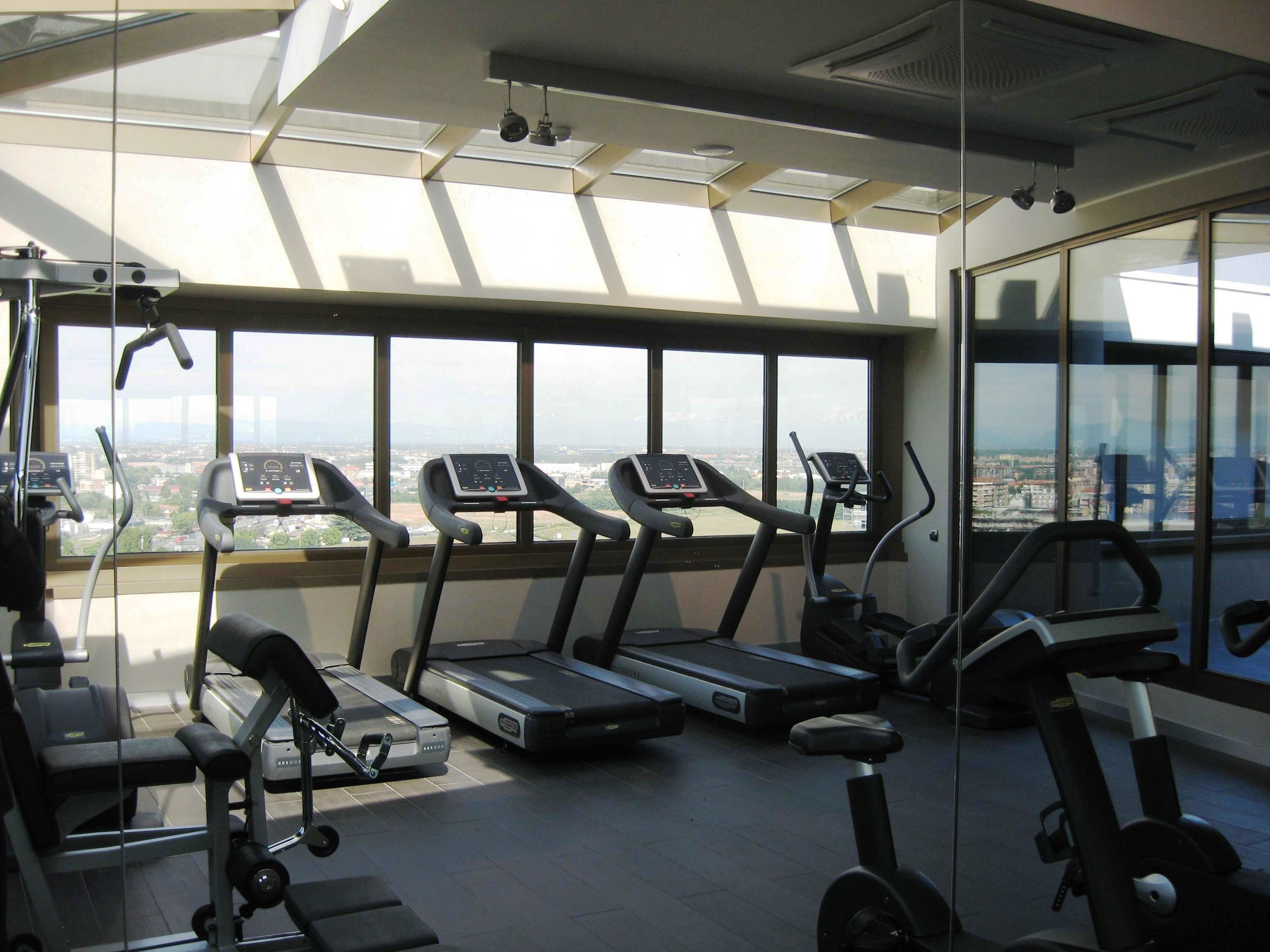 Un gimnasio con cintas de correr, bicicletas estáticas y una máquina de remo, con amplios ventanales que ofrecen vista a la ciudad.