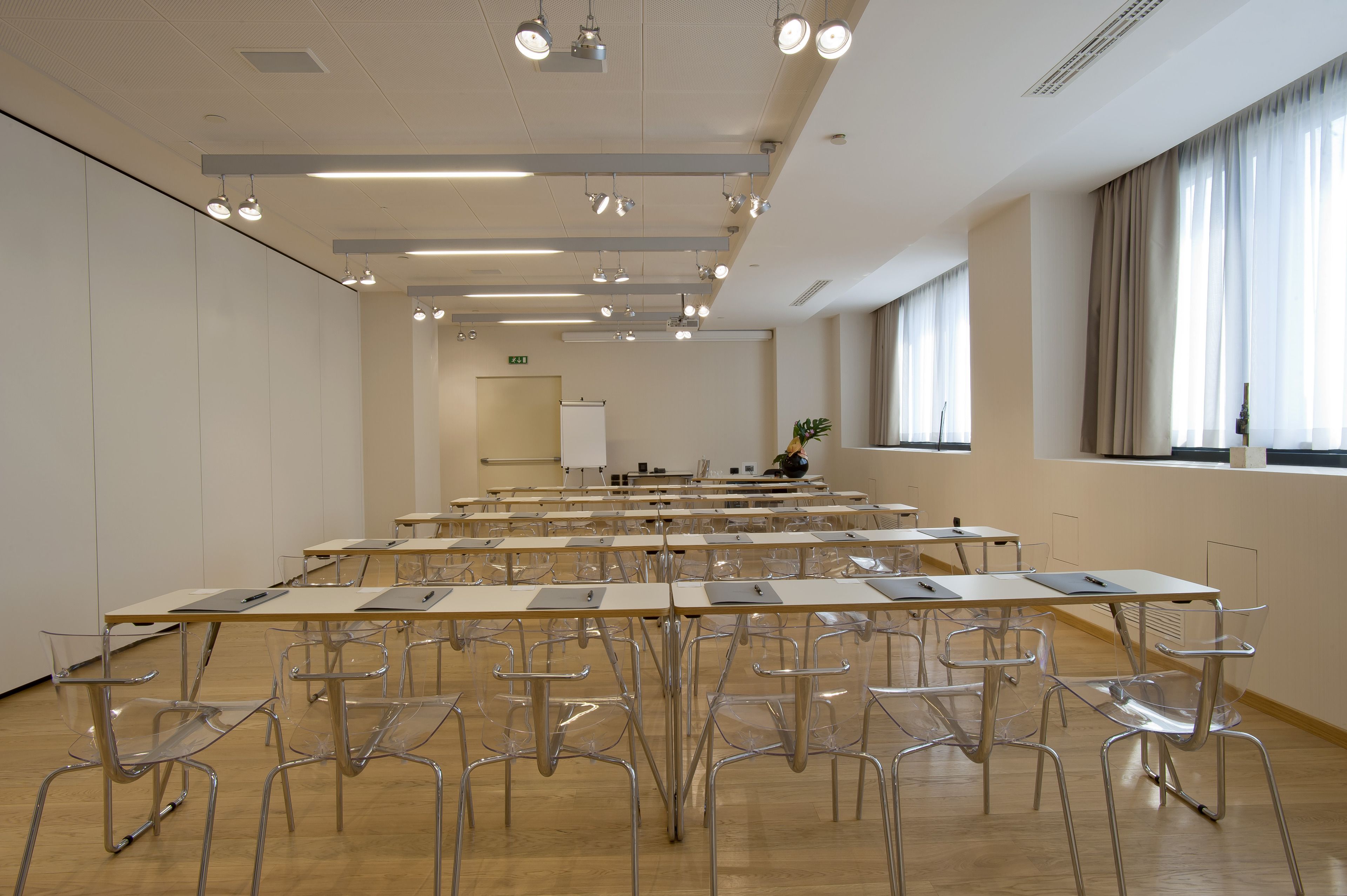 Uma sala de conferências ampla e bem iluminada, com fileiras de mesas e cadeiras, um quadro branco e uma planta perto das janelas.