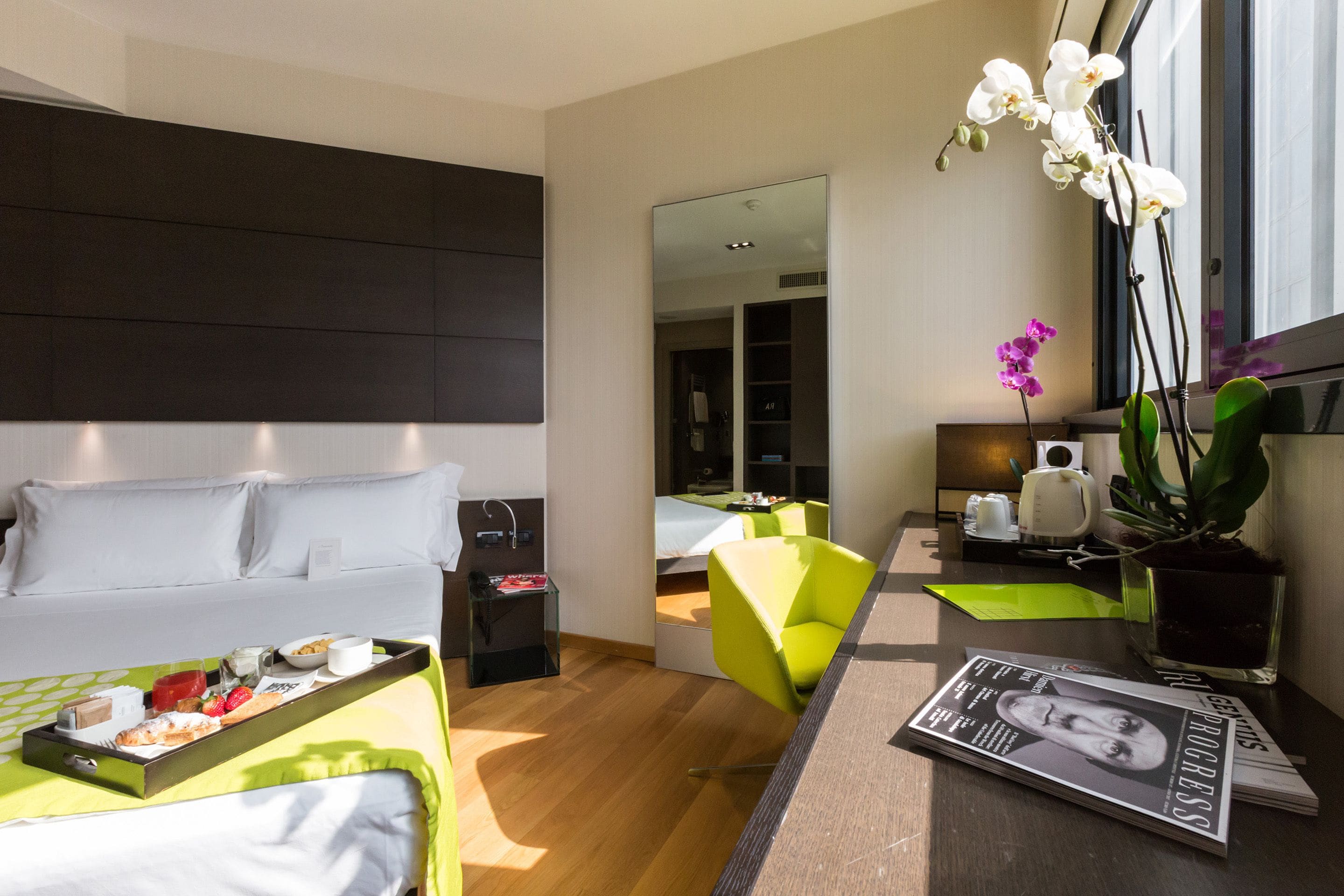 Habitació d'hotel moderna amb un llit, un escriptori amb una revista, una cadira verd brillant i una finestra gran amb orquídies.