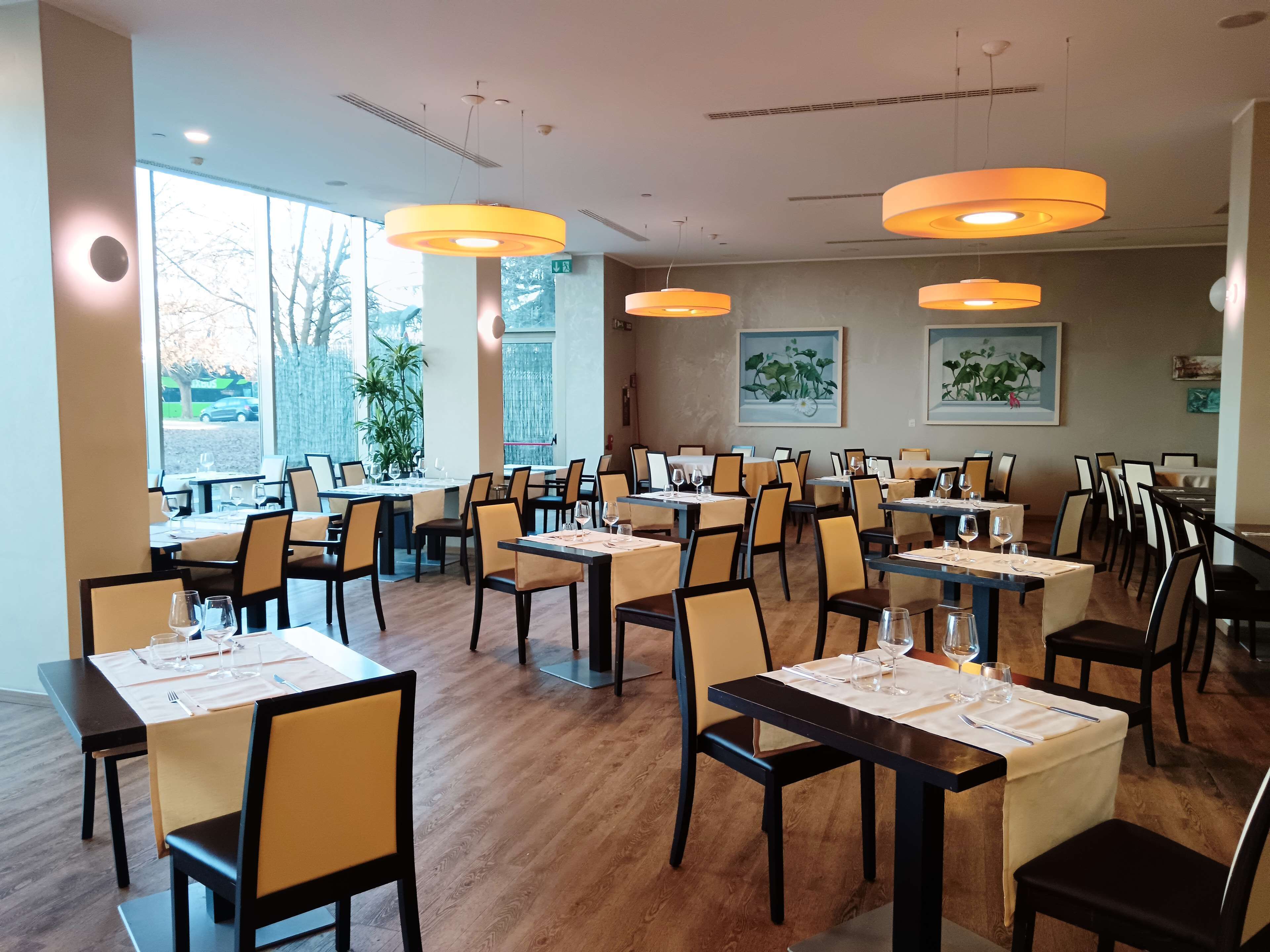 A restaurant with multiple tables and chairs, large windows, and hanging pendant lights.