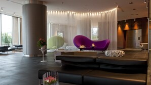Un hall d’entrée moderne avec un canapé noir, un fauteuil violet et un fauteuil vert. On y trouve une grande colonne et une table en verre sur laquelle est posé un vase de fleurs.