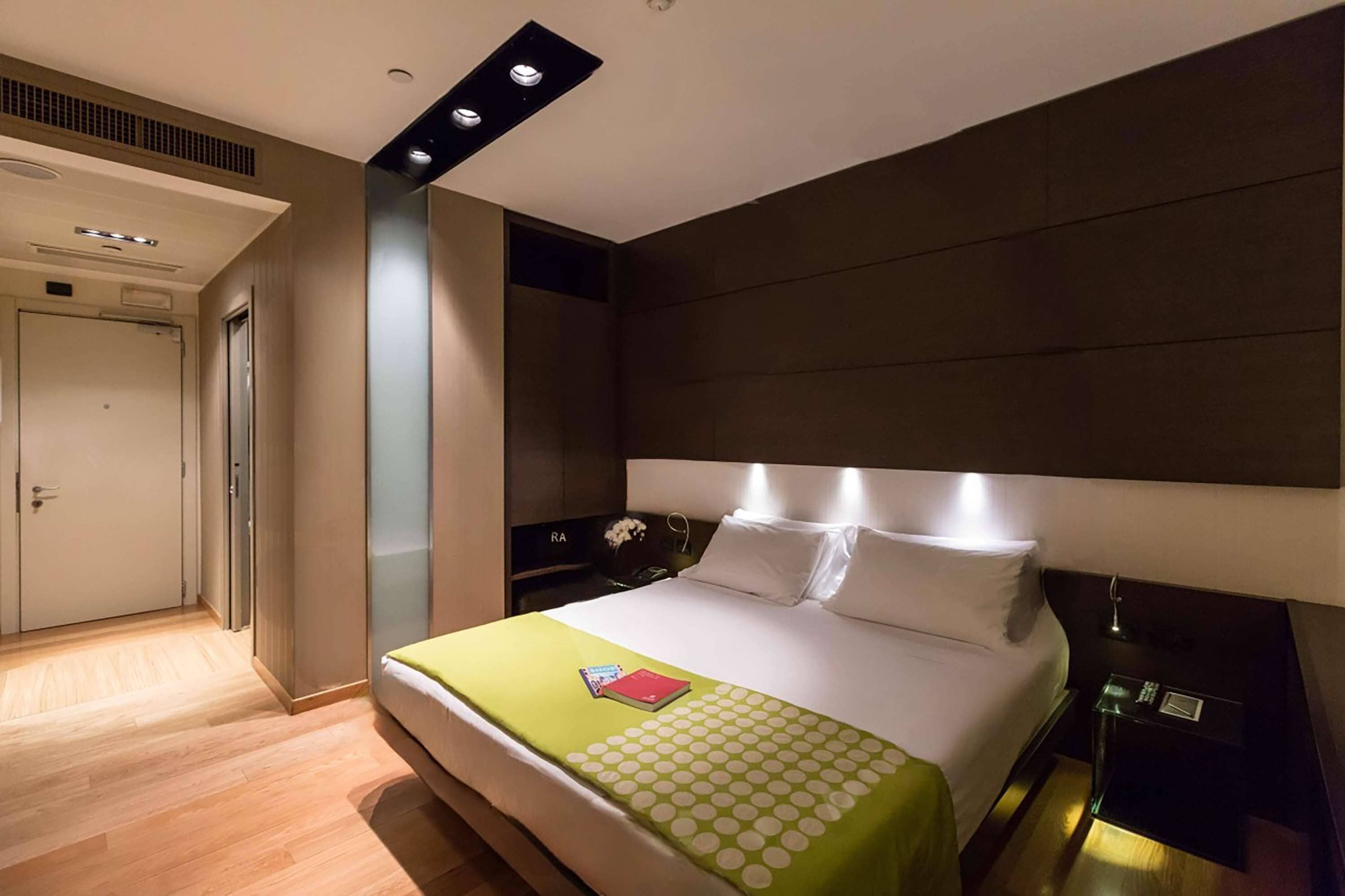 A modern hotel room with a large bed, a bedside table with a lamp, and a wardrobe.