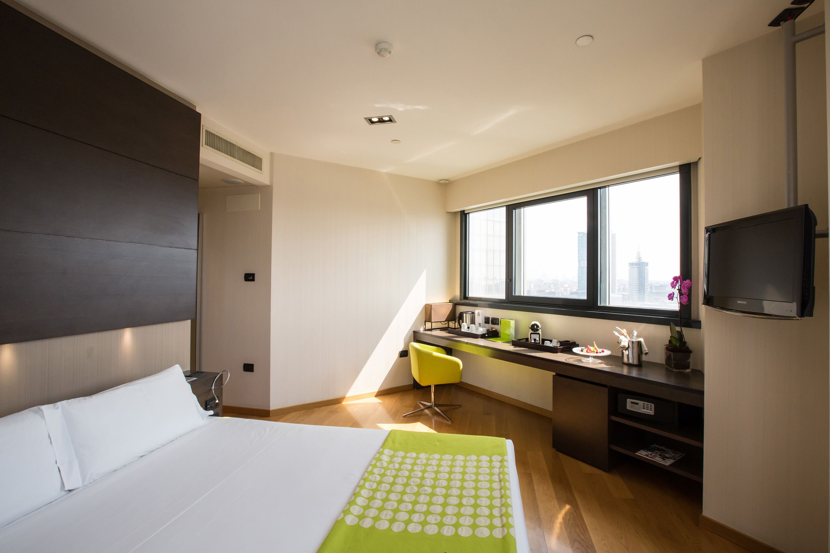 Ein modernes Hotelzimmer mit einem großen Bett, einem Schreibtisch mit Stuhl, einem Fernseher und einem Fenster mit Blick auf die Stadt.