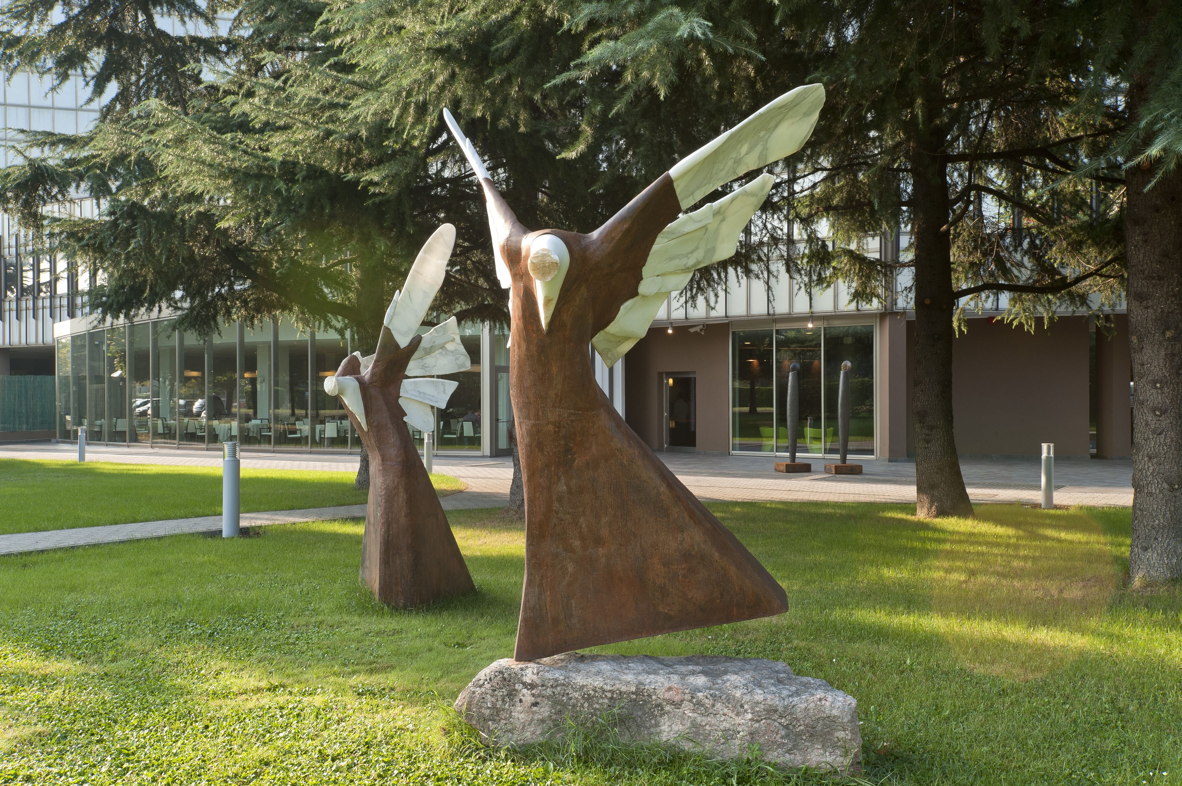 Escultura de un ave en vuelo sobre un césped con árboles y un edificio al fondo.
