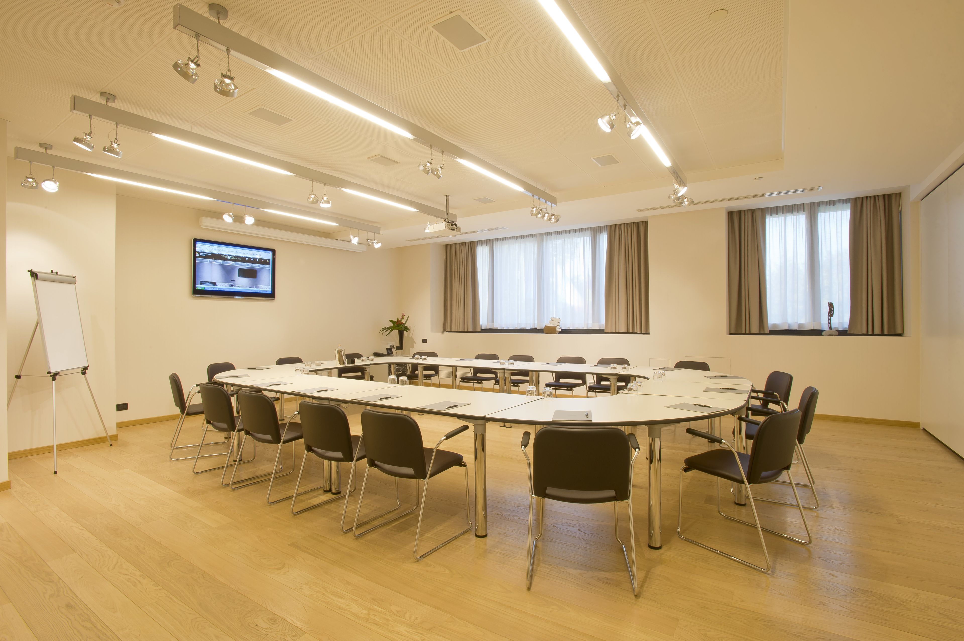 Un salón de conferencias con una larga mesa, sillas, una pizarra, una pantalla de televisión y ventanas grandes con cortinas.