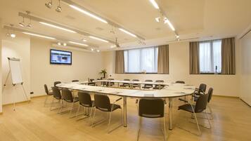 Un salón de conferencias con una larga mesa, sillas, una pizarra, una pantalla de televisión y ventanas grandes con cortinas.