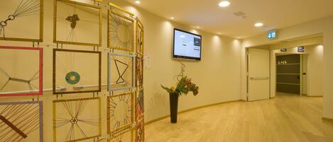 A hotel lobby with a wooden floor, a reception desk, and a wall-mounted screen displaying information.