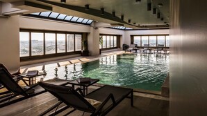 Une grande piscine intérieure dotée de nombreux chaises longues et de tables, de grandes fenêtres offrant une vue sur la ville et d’un design architectural moderne.