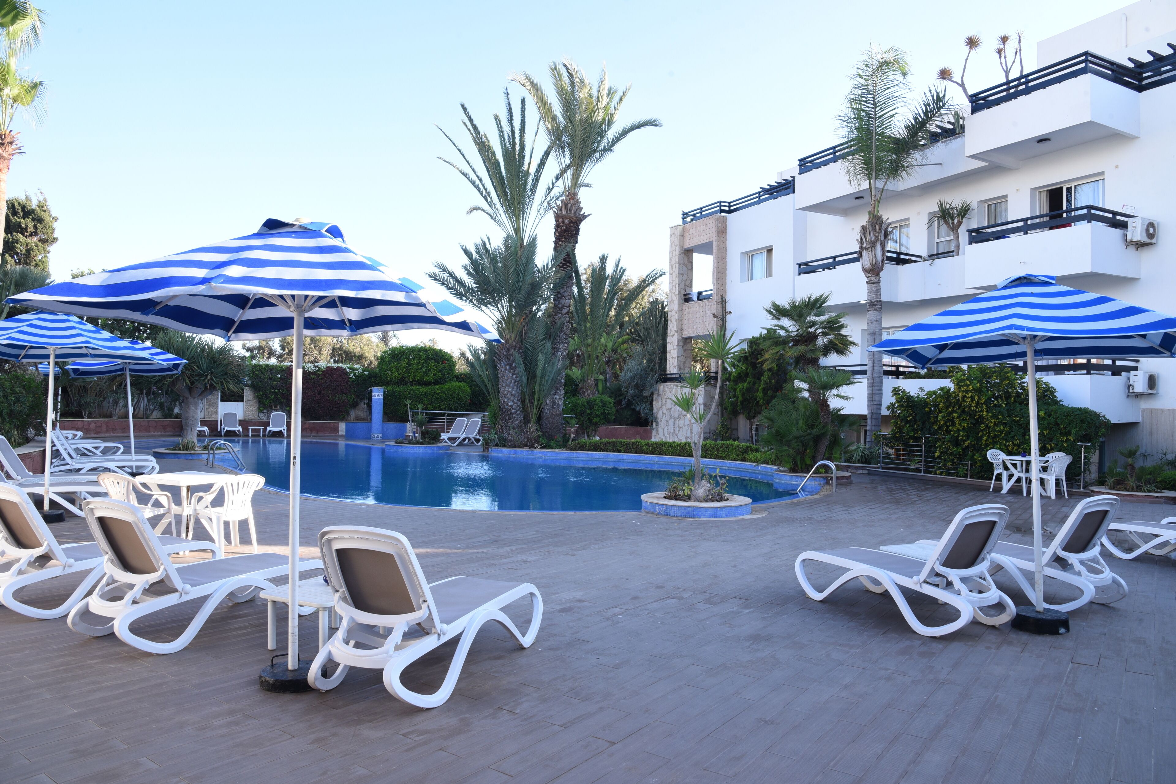 A poolside area with sun loungers and striped umbrellas, surrounded by palm trees and a building with balconies.
