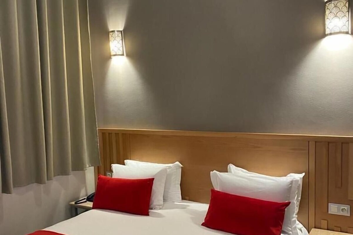 A hotel room with a large bed, two bedside tables, a nightstand, a wall-mounted lamp, and a window with curtains.