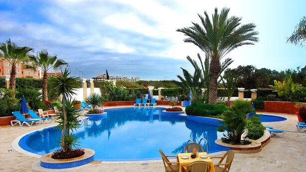 A swimming pool with lounge chairs and a table, surrounded by palm trees and a building in the background. - Golden Beach Appart'hotel (Agadir)