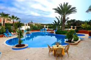 A swimming pool with lounge chairs and a table, surrounded by palm trees and a building in the background. - Golden Beach Appart'hotel (Agadir)