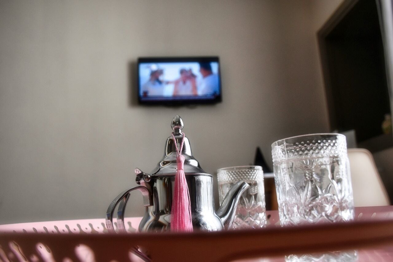 Eine silberne Teekanne mit einem rosafarbenen Quasten auf einer Tablett, ein Glas mit strukturierter Oberfläche und ein Fernseher, der eine Szene zeigt.