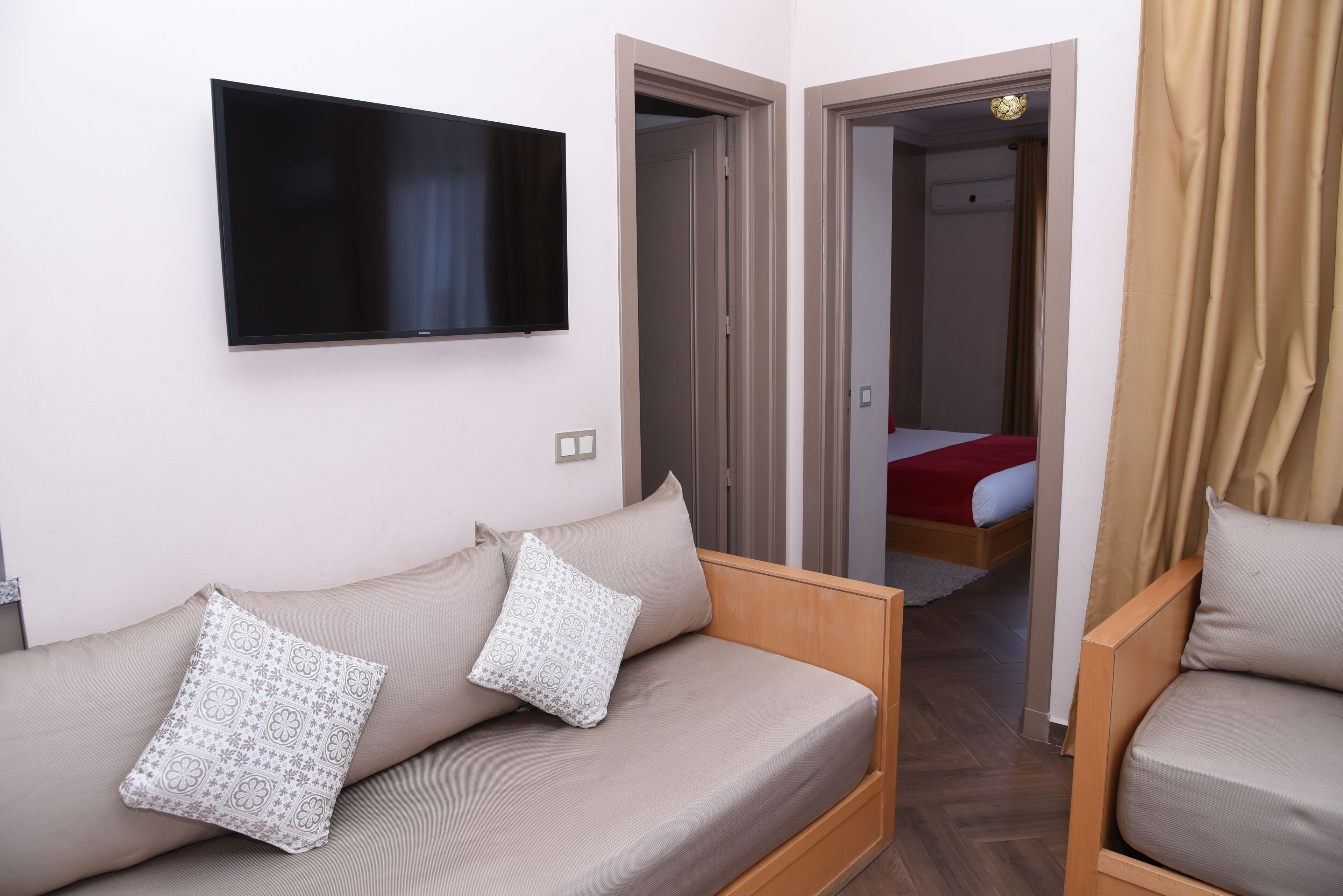 A modern hotel room with a flat-screen TV mounted on the wall, a sofa with patterned cushions, and a bed with a red blanket.