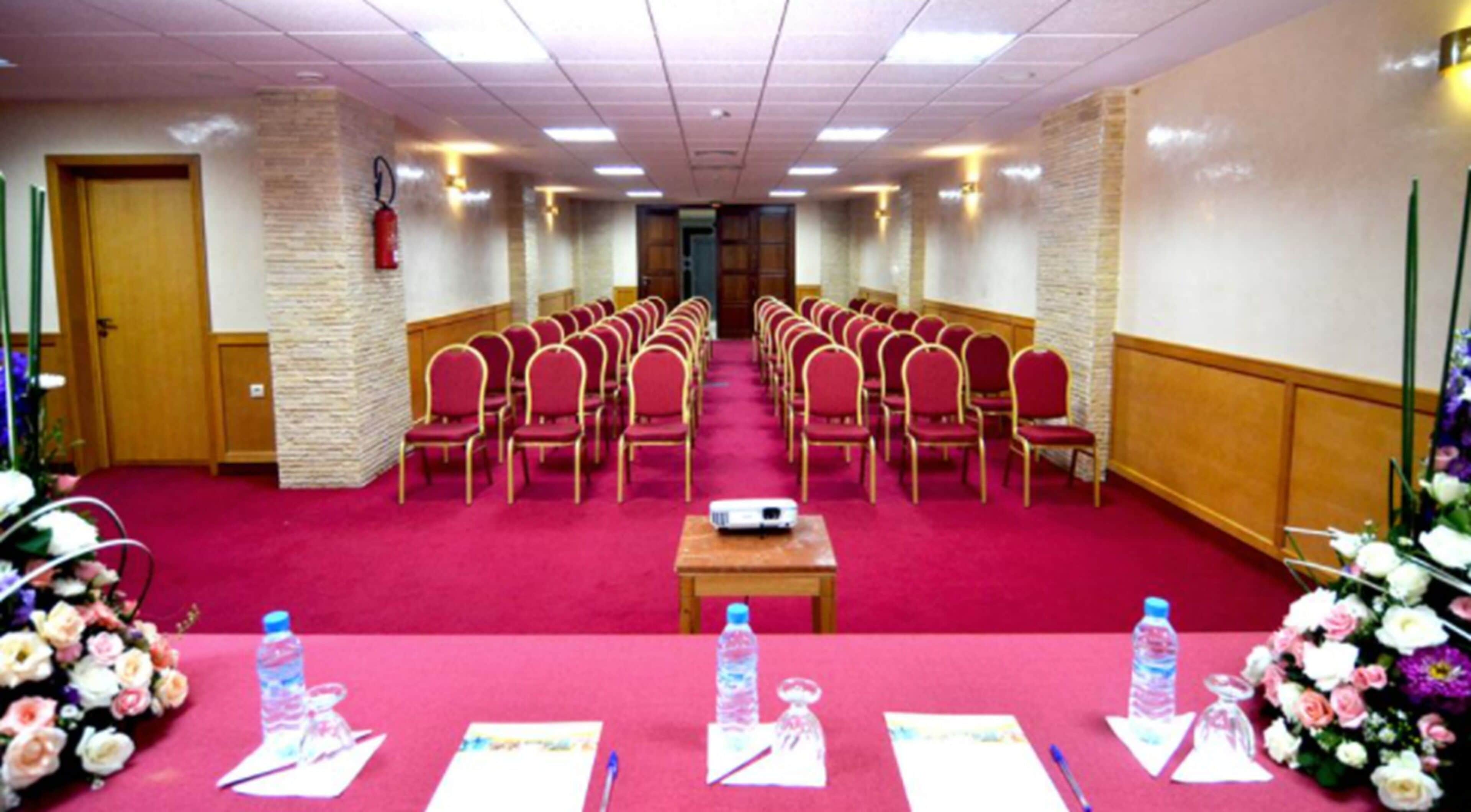 A conference room with rows of red chairs, a projector on a table, and floral arrangements.