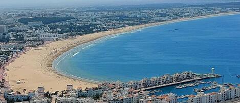 Un paisaje urbano costero con una playa extensa, un puerto deportivo con barcos y edificios residenciales.