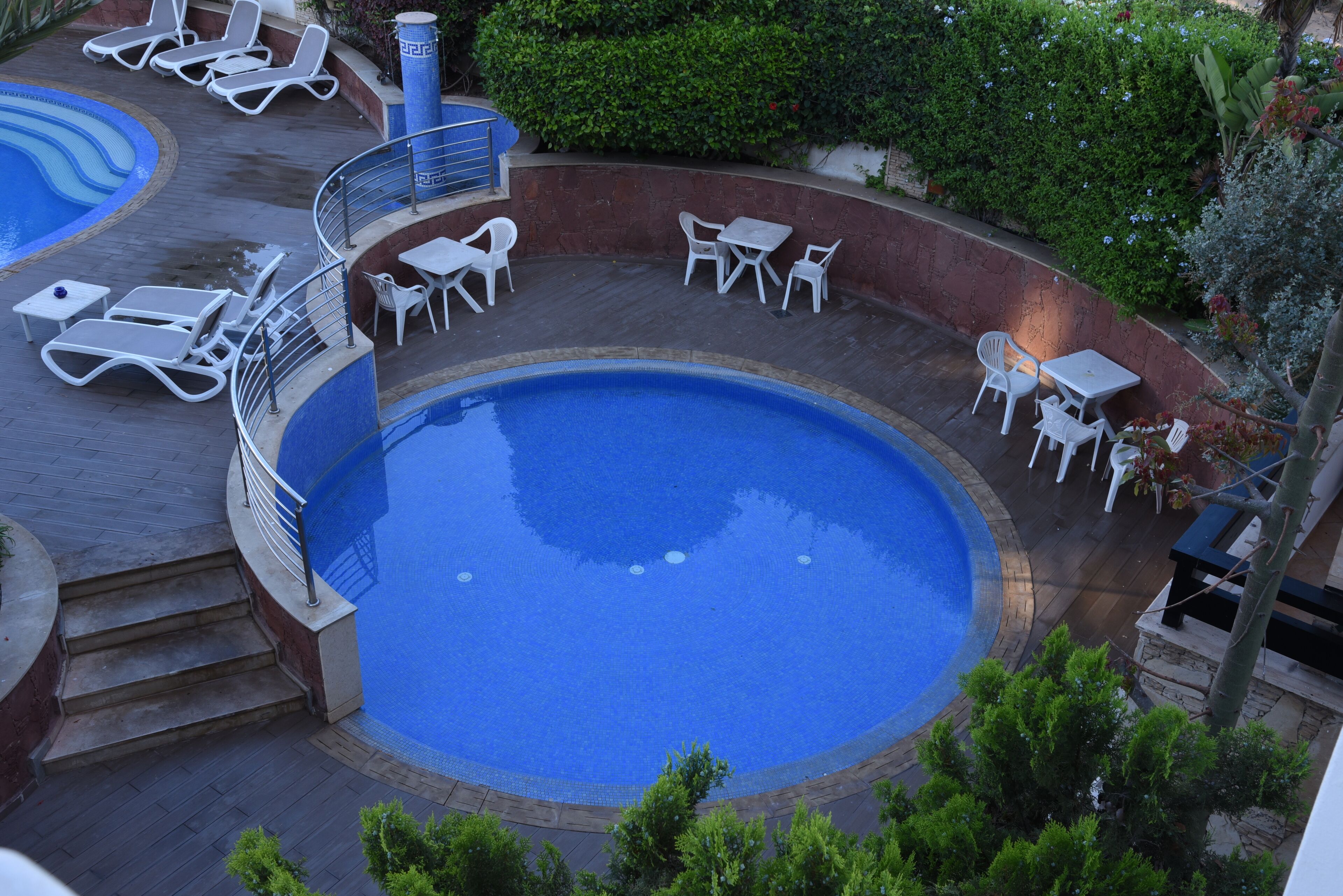 A circular pool with surrounding lounge chairs and tables, surrounded by a low brick wall and greenery.