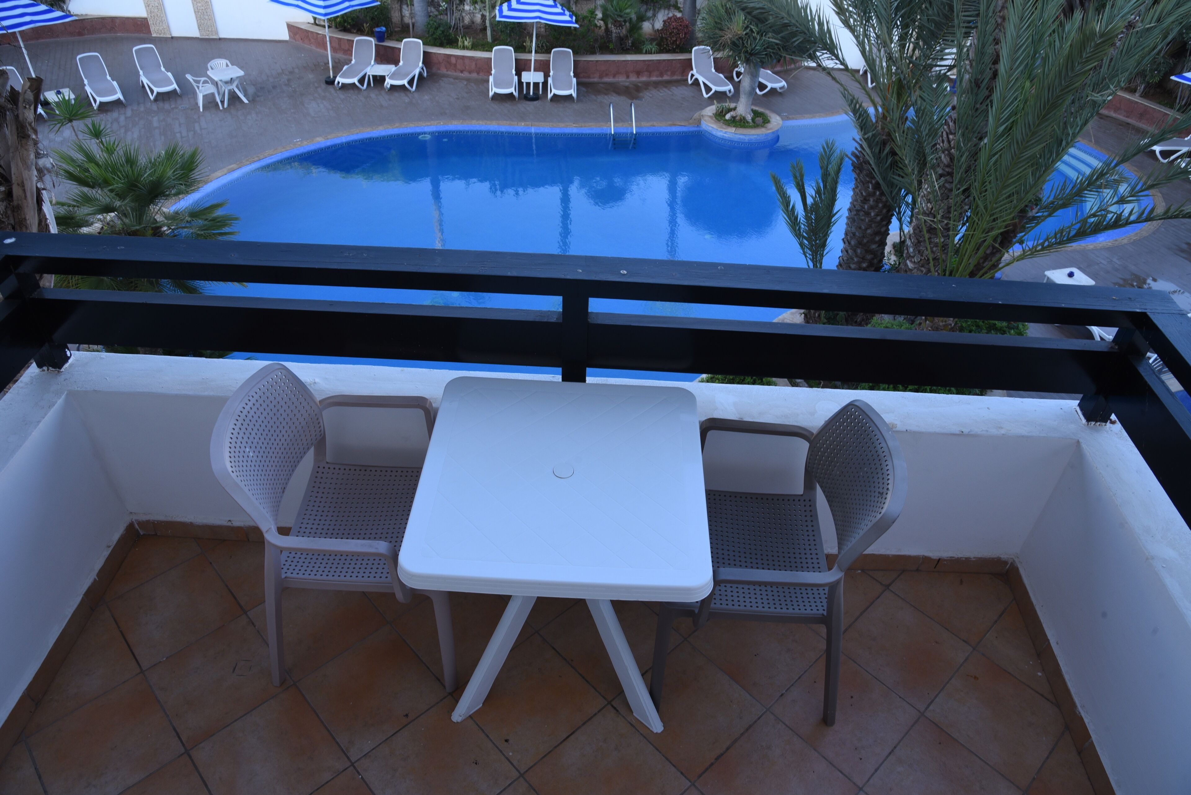 A balcony with two white plastic chairs and a small white table, overlooking a swimming pool with lounge chairs and umbrellas.