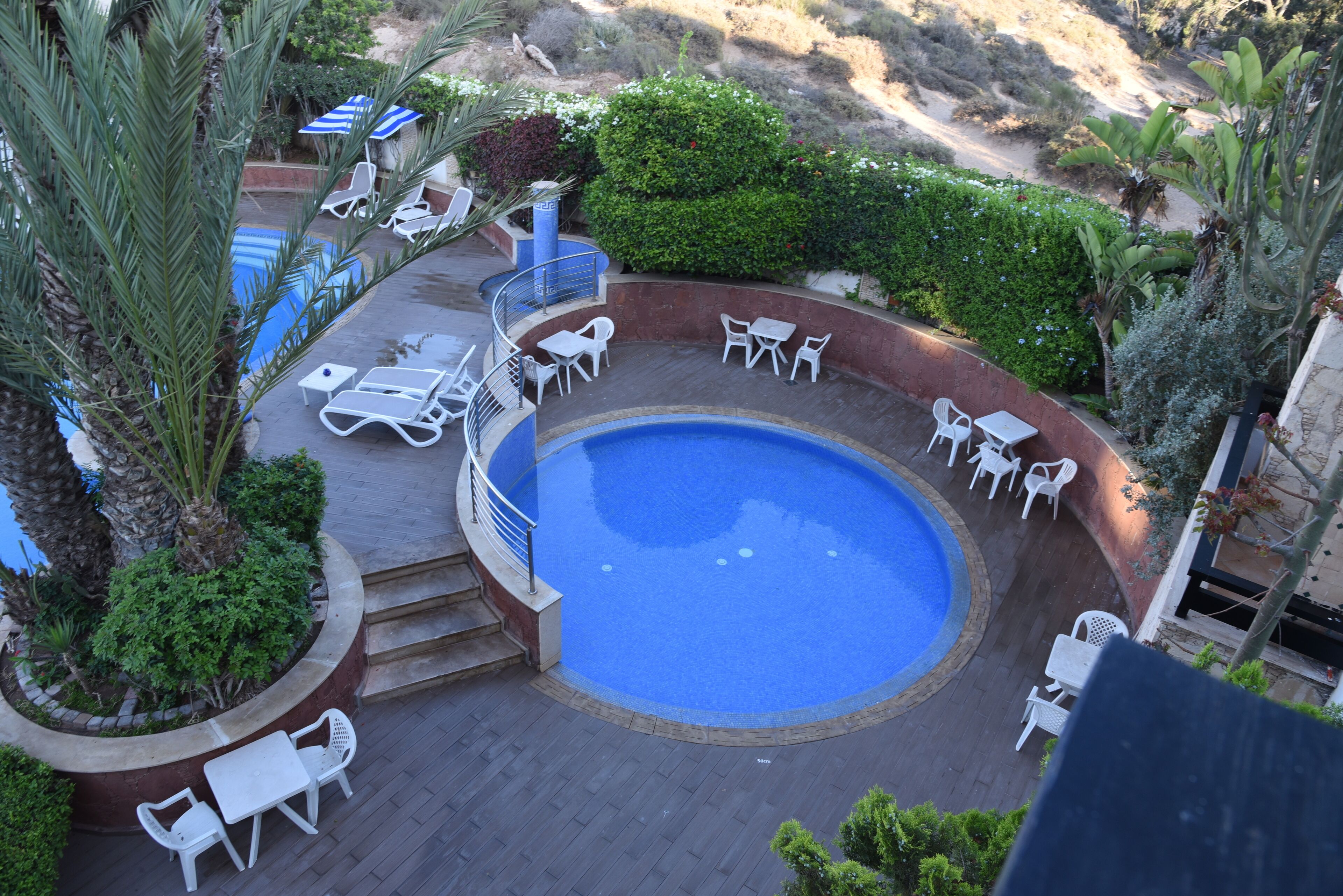 A circular pool with white lounge chairs and tables arranged around it, surrounded by greenery and a stone deck.