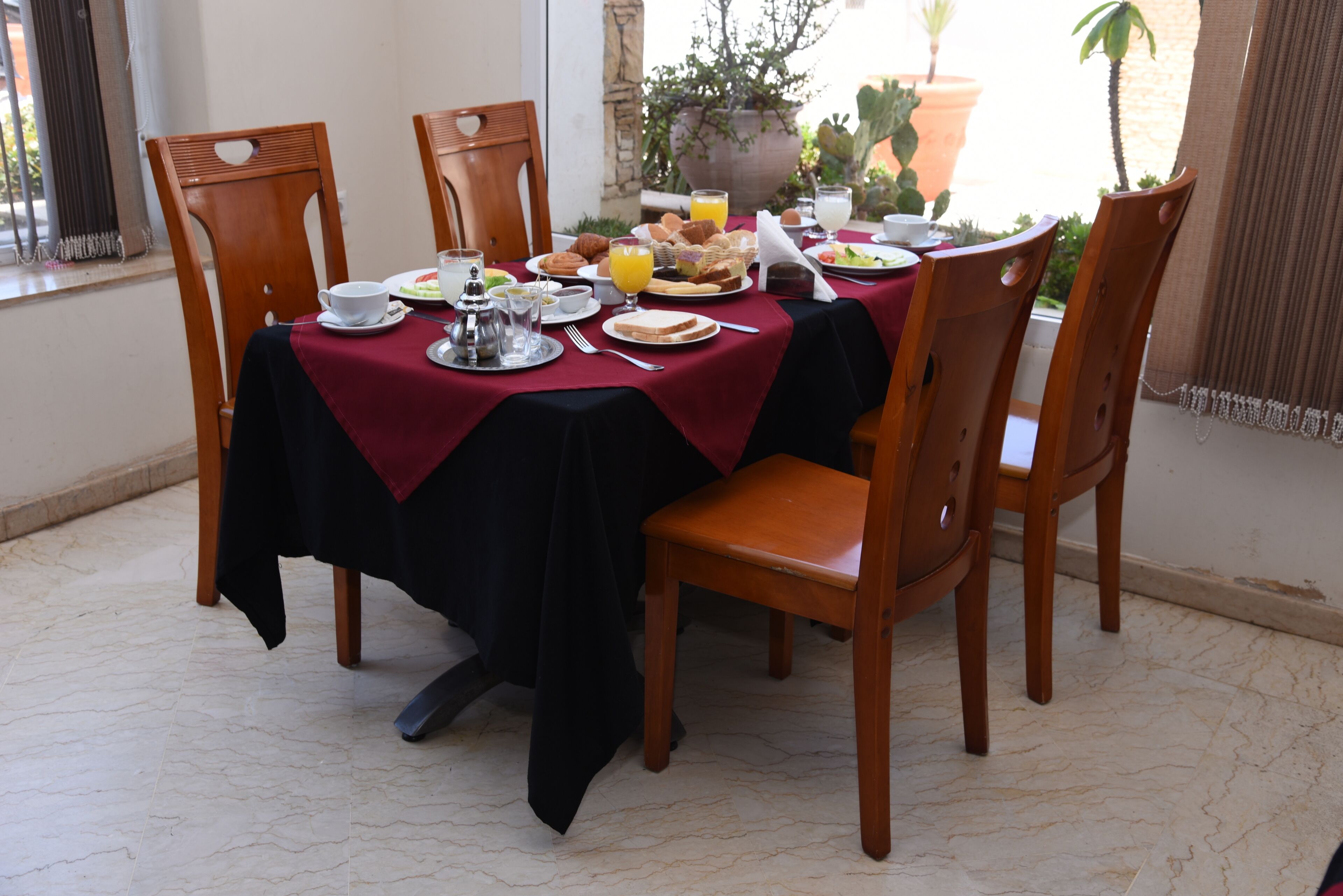 A dining table set for breakfast with a red tablecloth, wooden chairs, and various breakfast items.