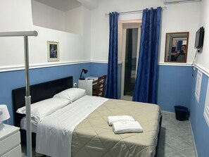 A bedroom with a bed, a desk, a chair, a bedside table, a lamp, a picture frame, a mirror, a trash bin, and blue curtains. - Hotel Alloggio Del Conte (Naples)
