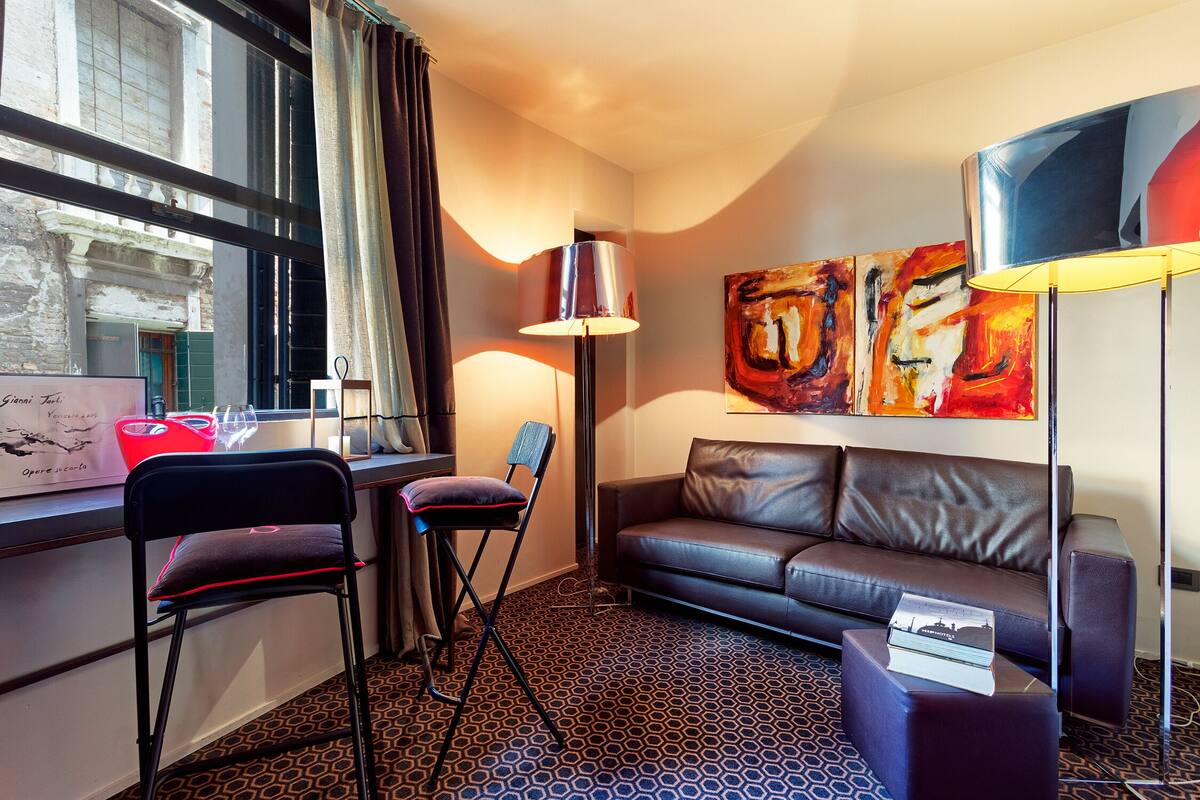 A modern hotel room with a leather sofa, a standing desk with a chair, a floor lamp, and a large abstract painting on the wall.