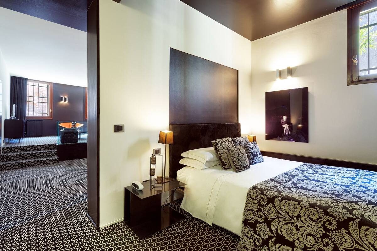 A hotel room with a large bed, a nightstand, a lamp, a painting on the wall, and a window with curtains.