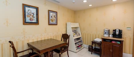 A hotel room with a dining table, chairs, a bookshelf, a cabinet with a coffee machine, and two framed pictures on the wall.