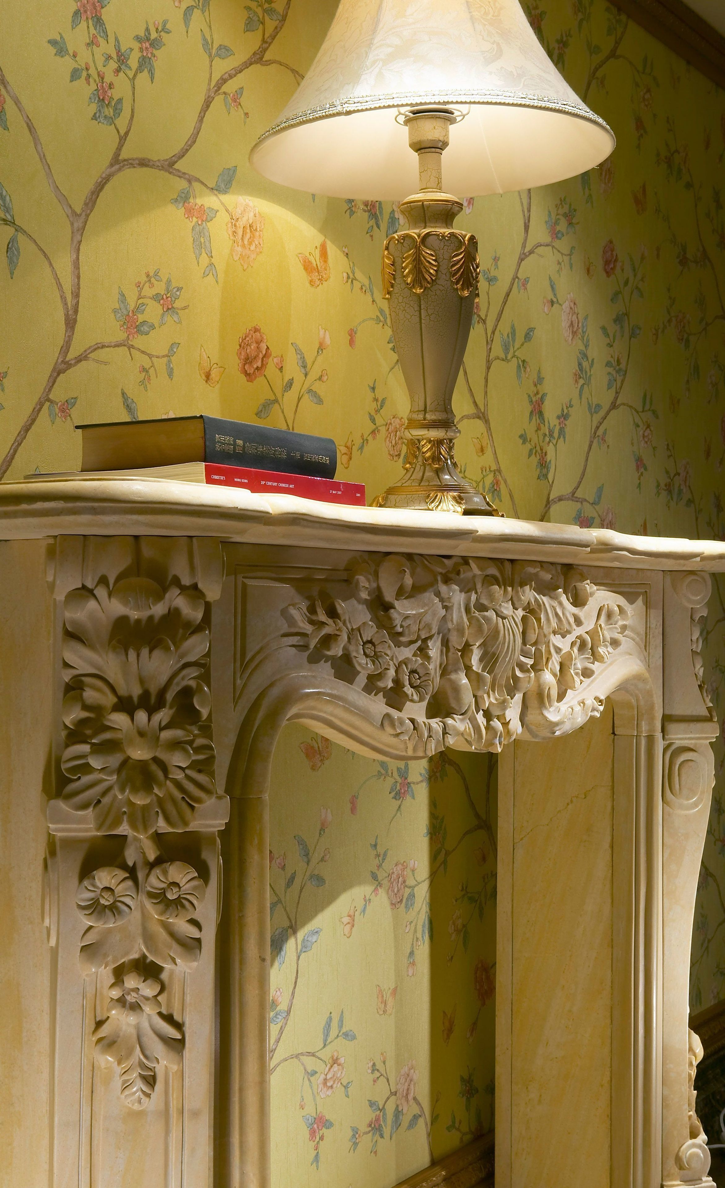 A marble fireplace with intricate floral carvings, a white lamp, and a book on top.