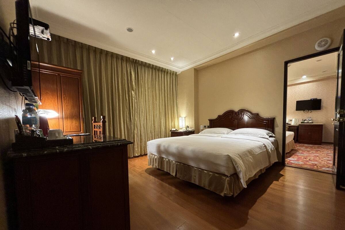 A hotel room with a large bed, a desk with a lamp, a television mounted on the wall, and a door leading to another room.