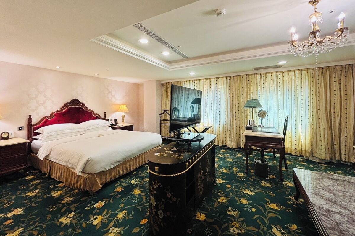 A hotel room with a large bed, a desk with a television, a chandelier, and a carpeted floor.