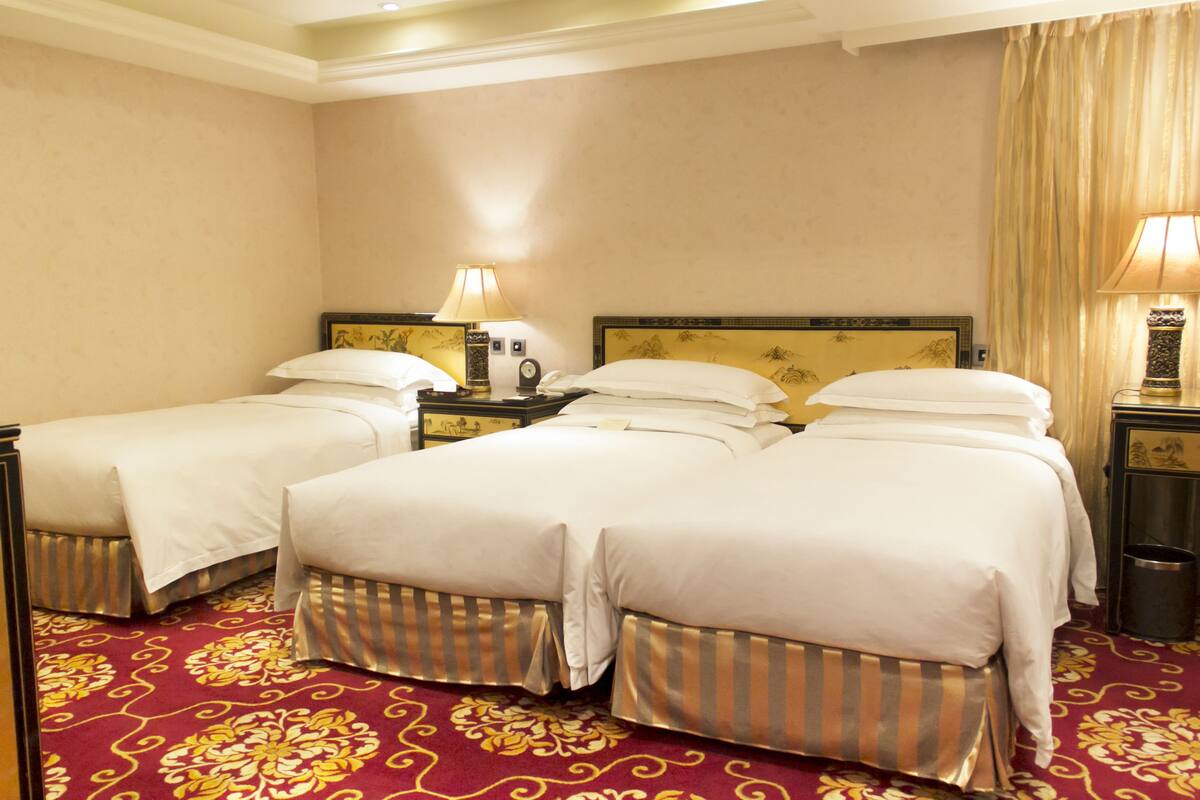 A hotel room with two single beds and a double bed, each with white linens and striped bed skirts. There are two bedside tables with lamps, a nightstand, and a trash can. The room has a red carpet with a floral pattern.