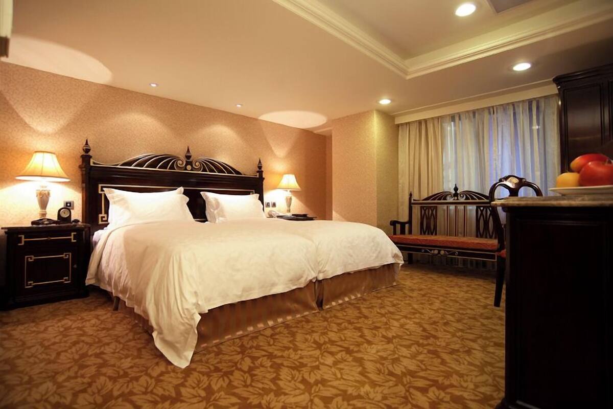 A hotel room with a large bed, two bedside tables with lamps, a chair, and a desk with fruit.