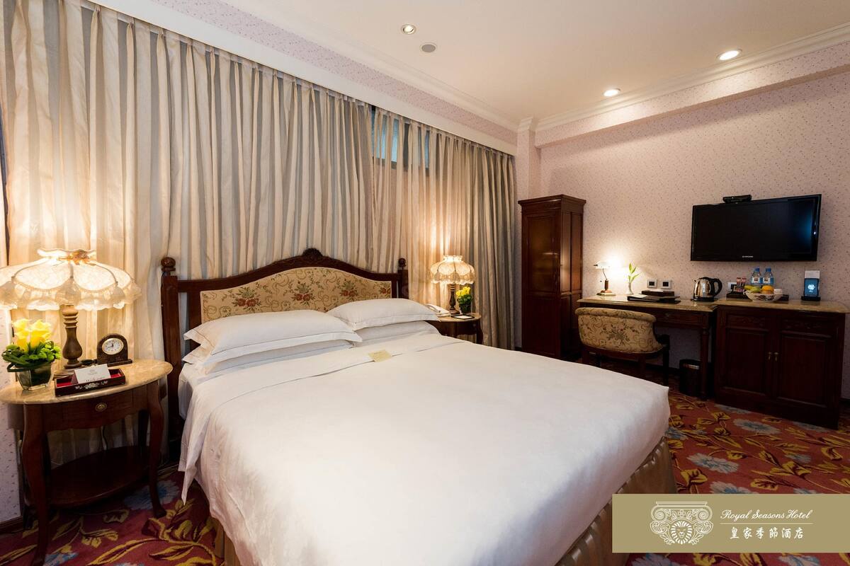 A hotel room with a large bed, two bedside tables with lamps, a desk with a chair, a television, and a wardrobe.