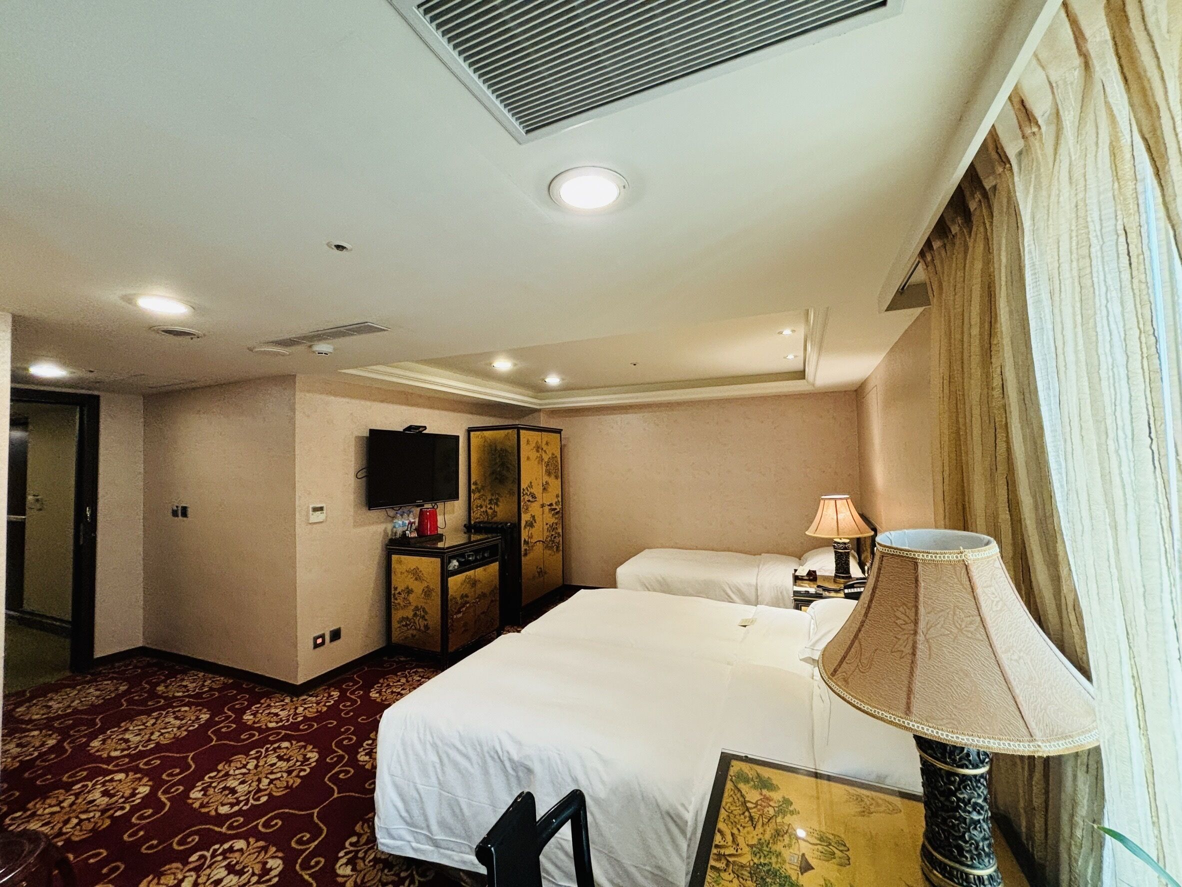 A hotel room with a bed, a desk with a lamp, a television, and a wardrobe.