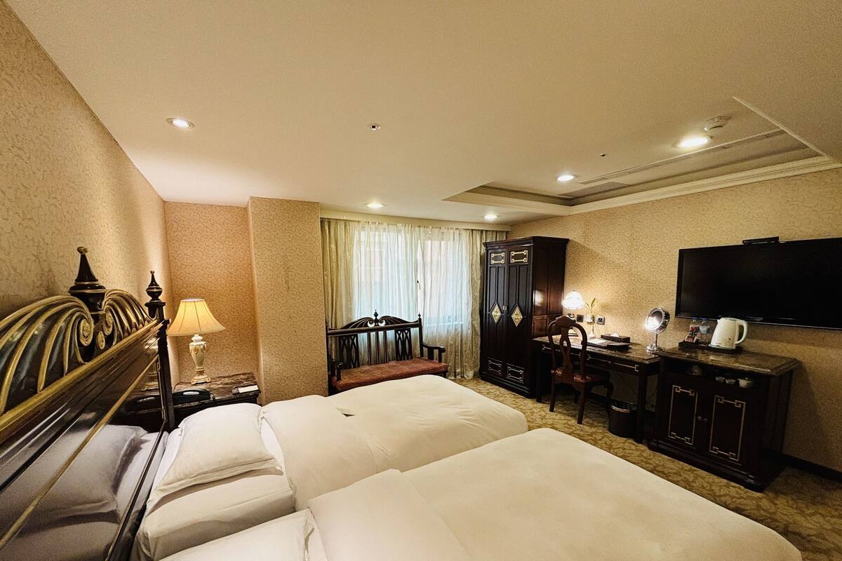 A hotel room with two beds, a desk, a chair, a wardrobe, a television, and a window with curtains.