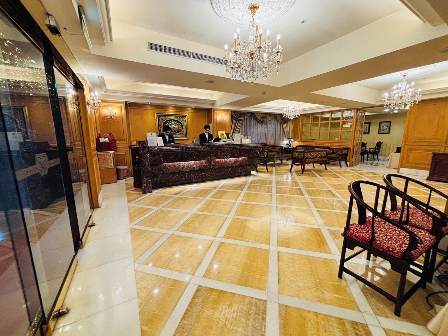 Un vestíbulo de hotel con un mostrador de recepción de mármol, paneles de madera, una araña y una zona de estar.