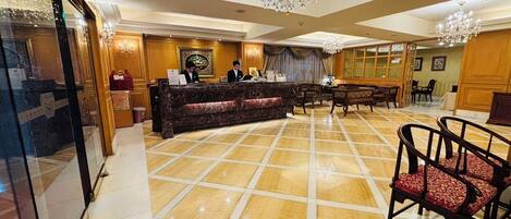 Un vestíbulo de hotel con un mostrador de recepción de mármol, paneles de madera, una araña y una zona de estar.