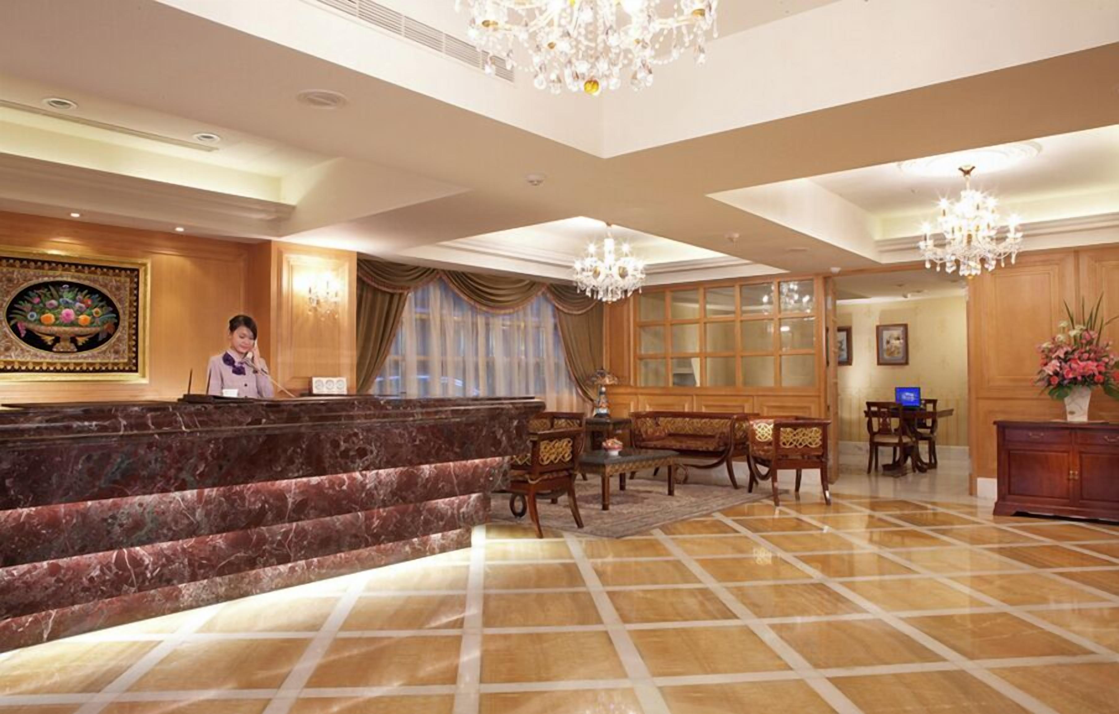 A hotel reception area with a marble reception desk, wooden furniture, chandeliers, and a large window with curtains.