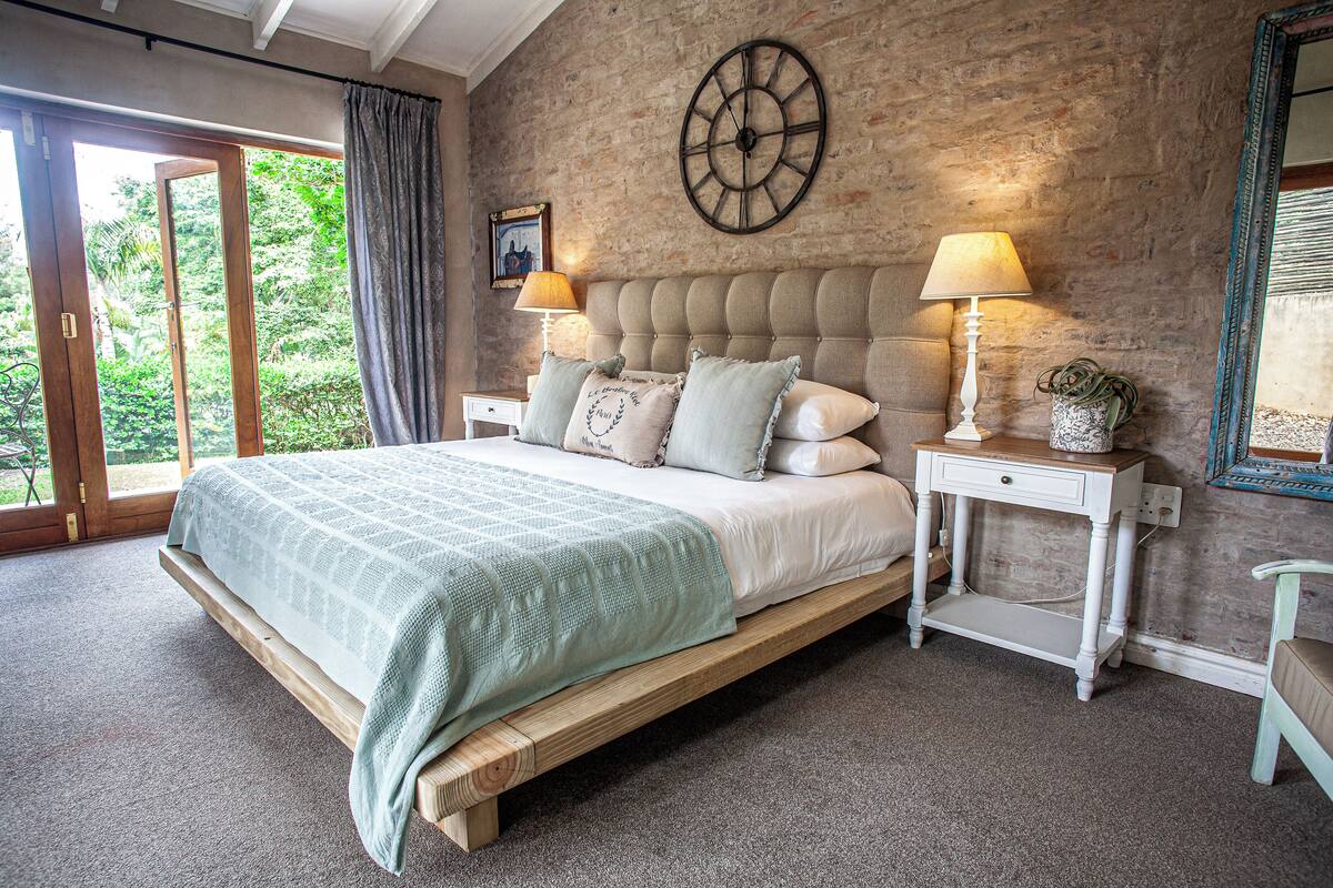 A bedroom with a large bed, two bedside tables with lamps, a wall clock, and a view of greenery through a sliding door.