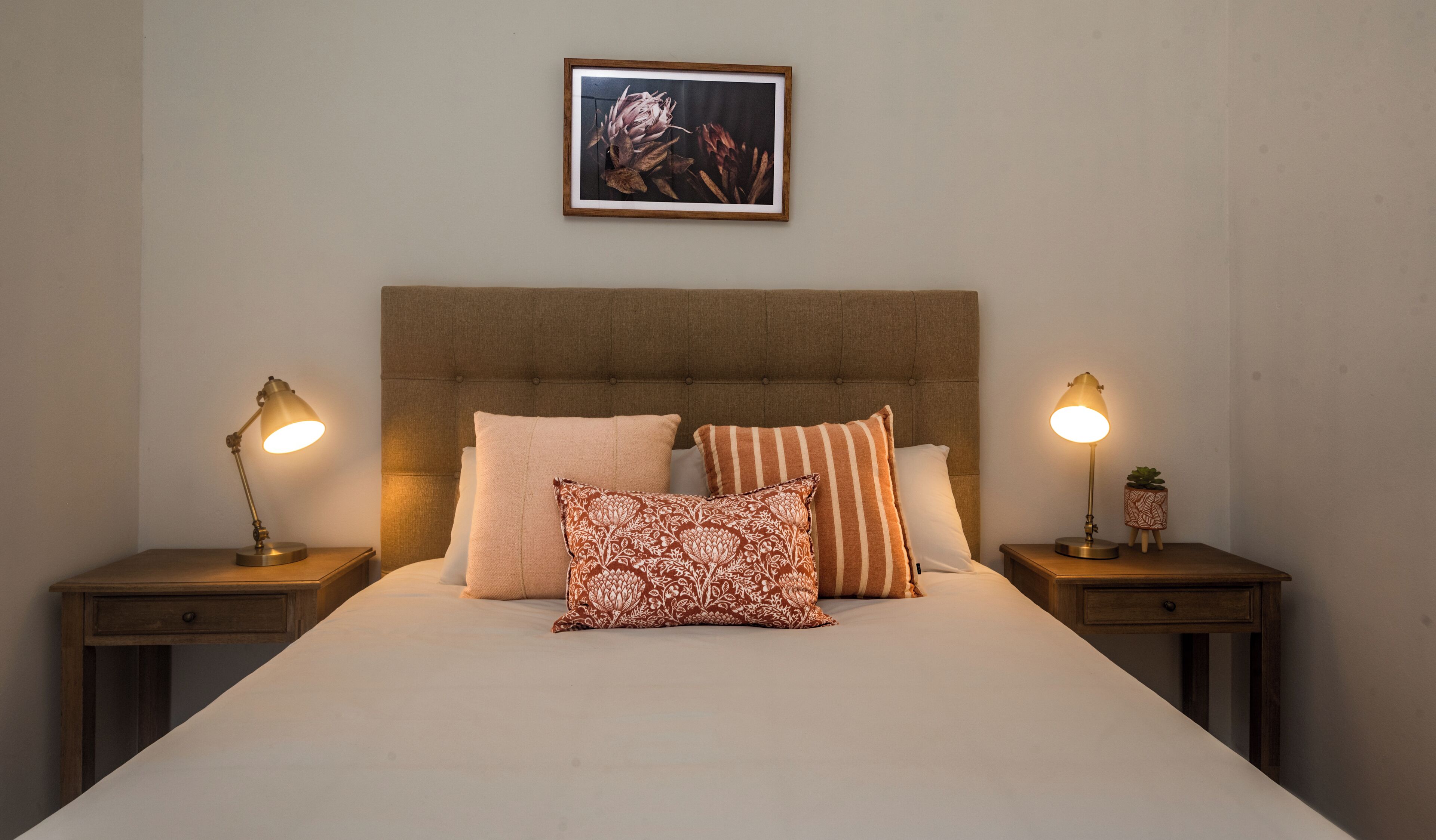 Una cama bien tendida con cabecera capitonada, dos lámparas de noche y un cuadro enmarcado sobre la cama.