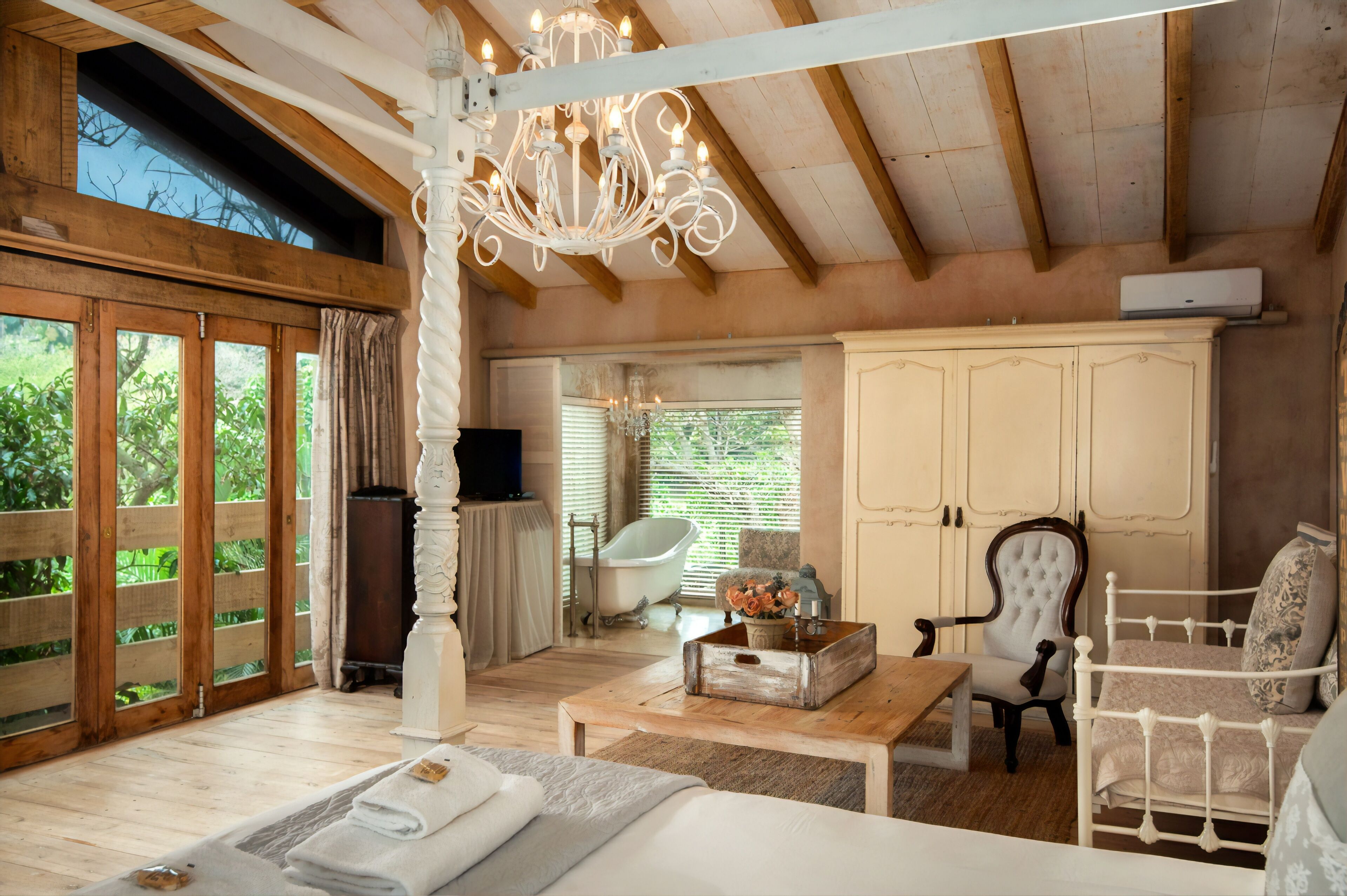 A spacious room with a wooden ceiling, a chandelier, a bathtub, a white wardrobe, and a comfortable seating area.