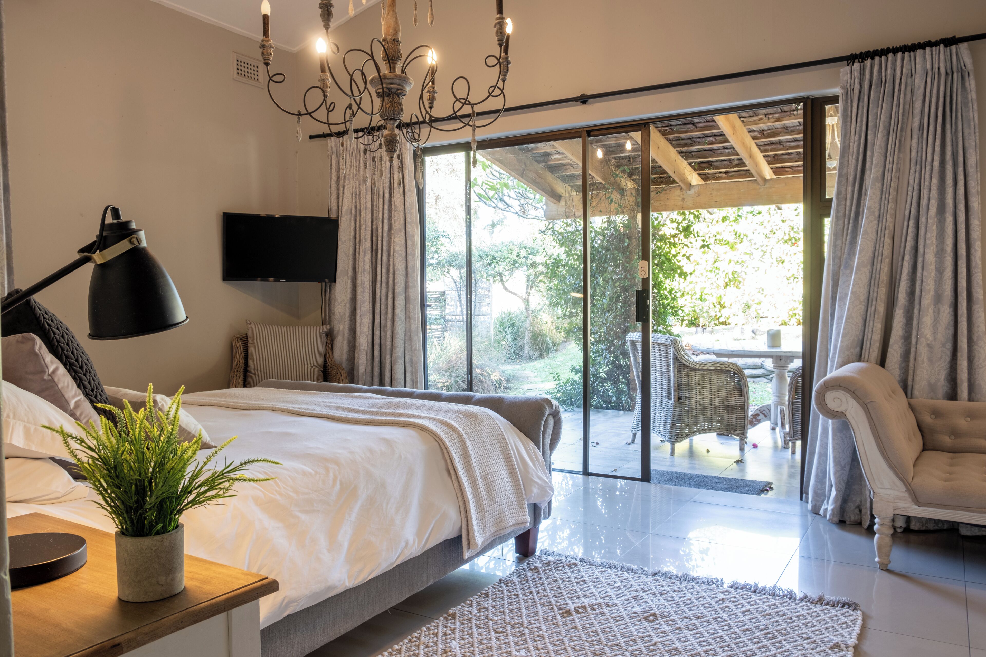 Quarto com cama, luminária de cabeceira, planta em vaso, televisão e porta de vidro deslizante que dá para área externa com móveis de vime.