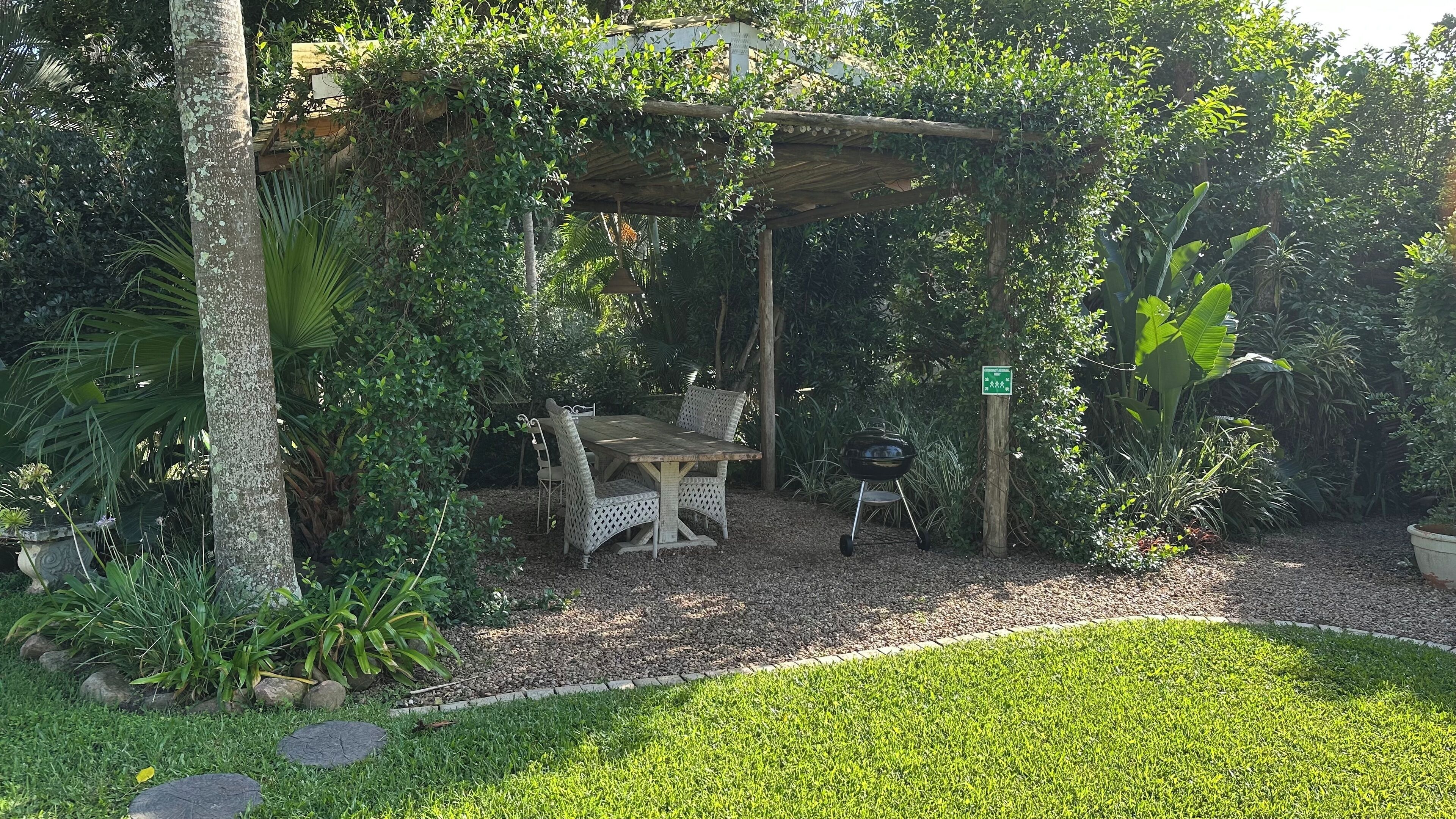 Un patio al aire libre con mesa y sillas de madera, rodeado de abundante follaje y una pérgola.