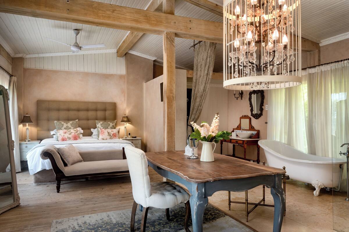 A bedroom with a large bed, a wooden table, a chandelier, a bathtub, and a nightstand with a lamp.