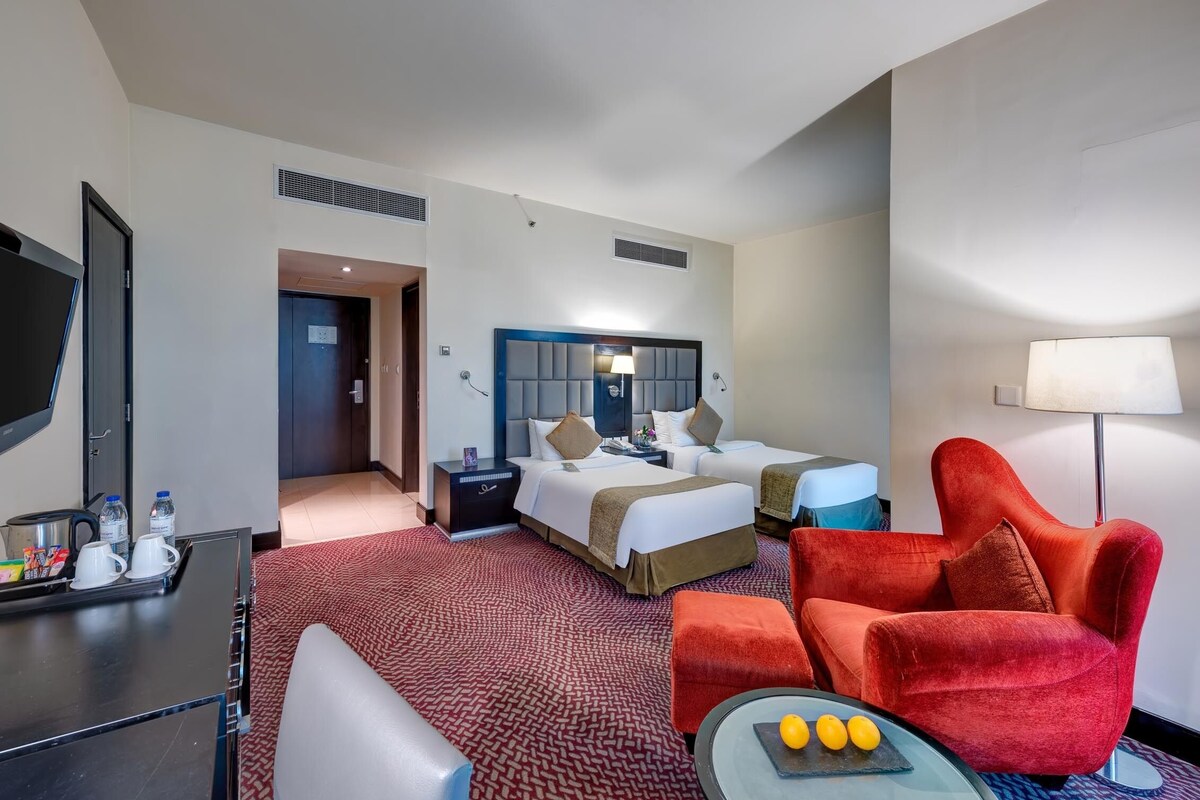 A hotel room with a bed, two bedside tables, a TV, a red armchair, and a small table with fruit.